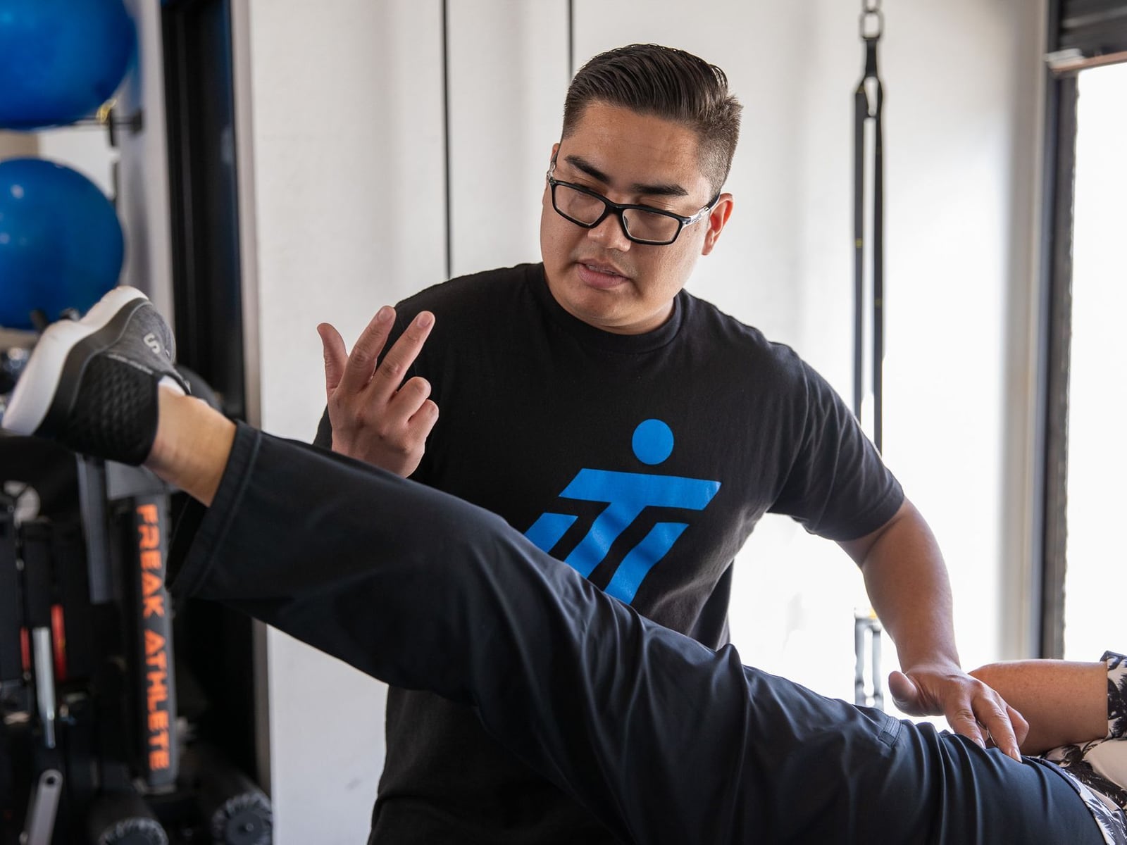 An IMTI practitioner in a logo tee assessing a client's hip and hamstring range, one hand steadying the raised leg